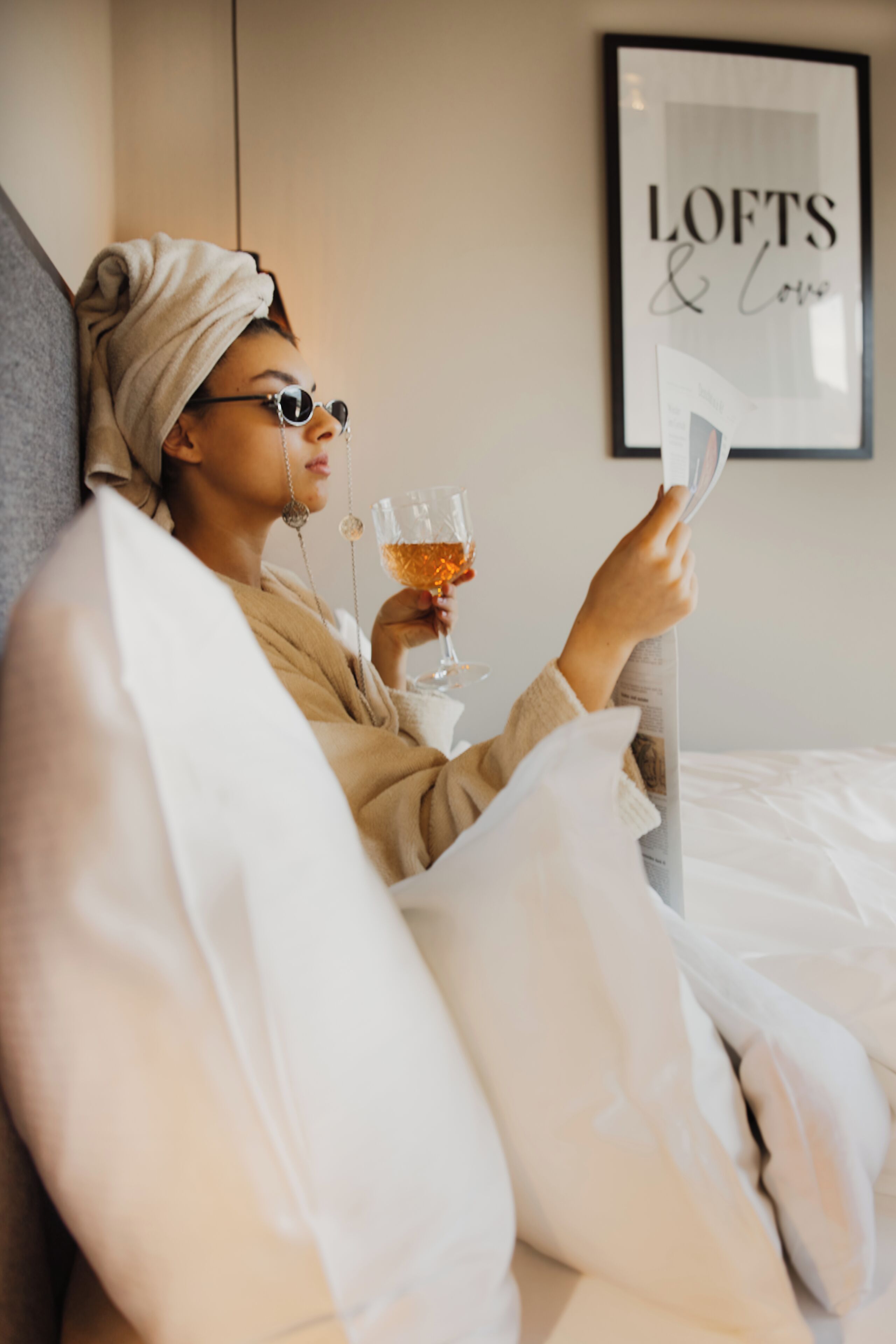 Una persona recostada en una cama con una almohada, sosteniendo un vaso con líquido ámbar y leyendo una revista. En la pared se ve un cartel enmarcado con el texto 'LOFTS & Love'.