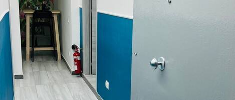 A hallway with a blue and white wall, a door with a plaque, a fire extinguisher, and a small table with a chair.