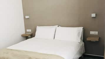 Cama bem arrumada com lençóis brancos e manta bege, posicionada contra uma parede em tons neutros com duas mesas de cabeceira.