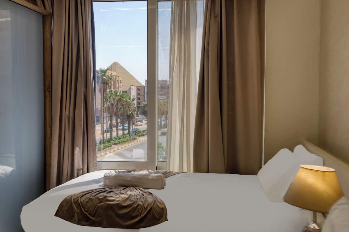 A hotel room with a bed, a window with curtains, a lamp, and a view of a pyramid and palm trees outside.