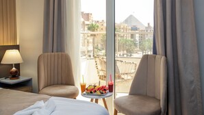 A hotel room with a bed, two armchairs, a small table with a tray of fruit and drinks, and a view of buildings and a pyramid through the window.