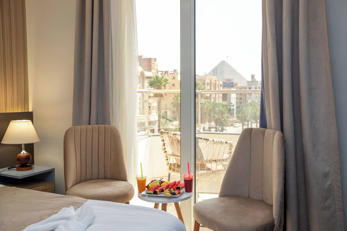 A hotel room with a bed, two armchairs, a small table with a tray of fruit and drinks, and a view of buildings and a pyramid through the window.
