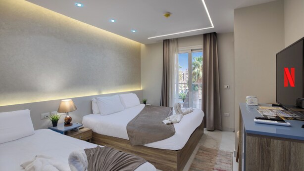 A hotel room with two beds, a desk with a TV, and a view of palm trees through the window.