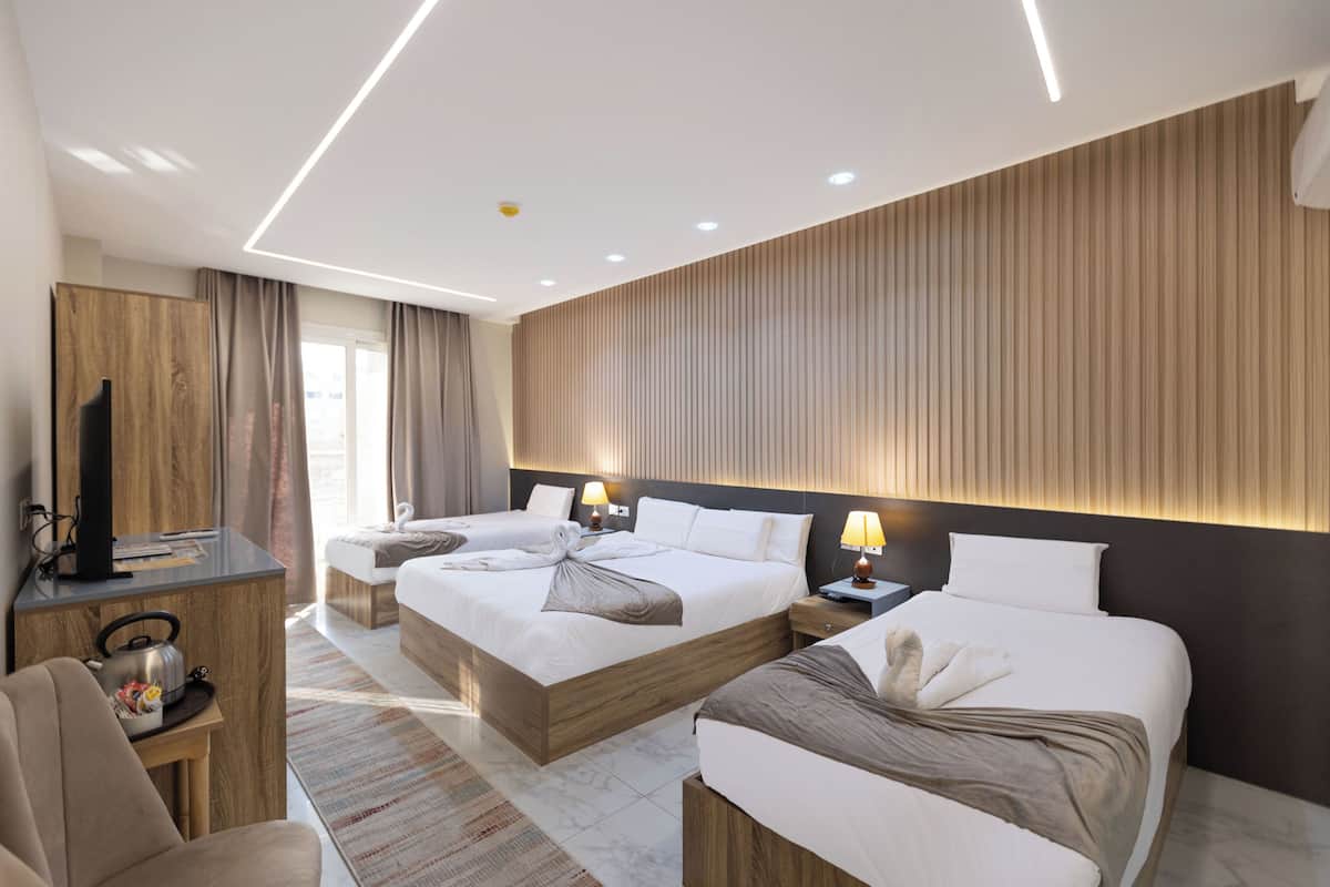 A modern hotel room with three beds, a desk with a television, and a large window.