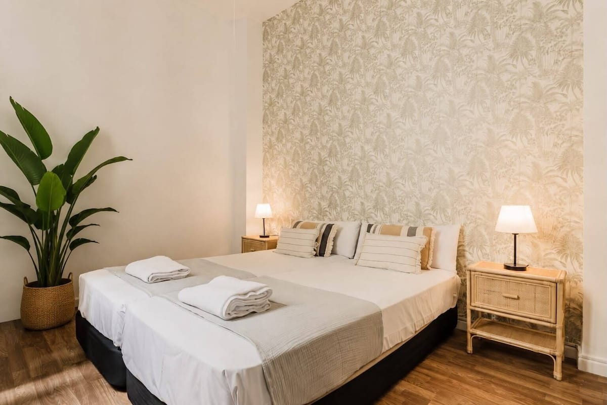 Een netjes opgemaakt bed met kussens, een bedlampje, een potplant en behang met een patroon.