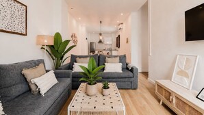 Un salon moderne avec deux canapés gris, une table basse agrémentée de plantes, un téléviseur et une cuisine en arrière-plan.