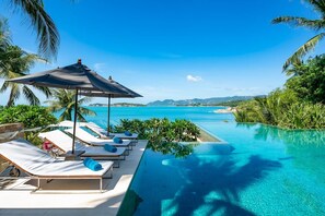 Ein Poolbereich mit Liegestühlen und Sonnenschirmen mit Blick auf das klare, blaue Meer und üppiges Grün.