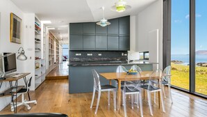 A modern kitchen with dark cabinets, a wooden dining table with clear chairs, and a view of the sea. - Tuath na Mara - Three Bedroom Villa, Sleeps 9 (Doagh Beg)