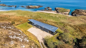 A modern building with a flat roof, situated on a grassy hill overlooking a coastal area with rocky outcrops and a clear blue sea. - Tuath na Mara - Three Bedroom Villa, Sleeps 9 (Doagh Beg)