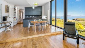 A modern kitchen with a view of the sea, wooden flooring, and a dining area with clear chairs. - Tuath na Mara - Three Bedroom Villa, Sleeps 9 (Doagh Beg)
