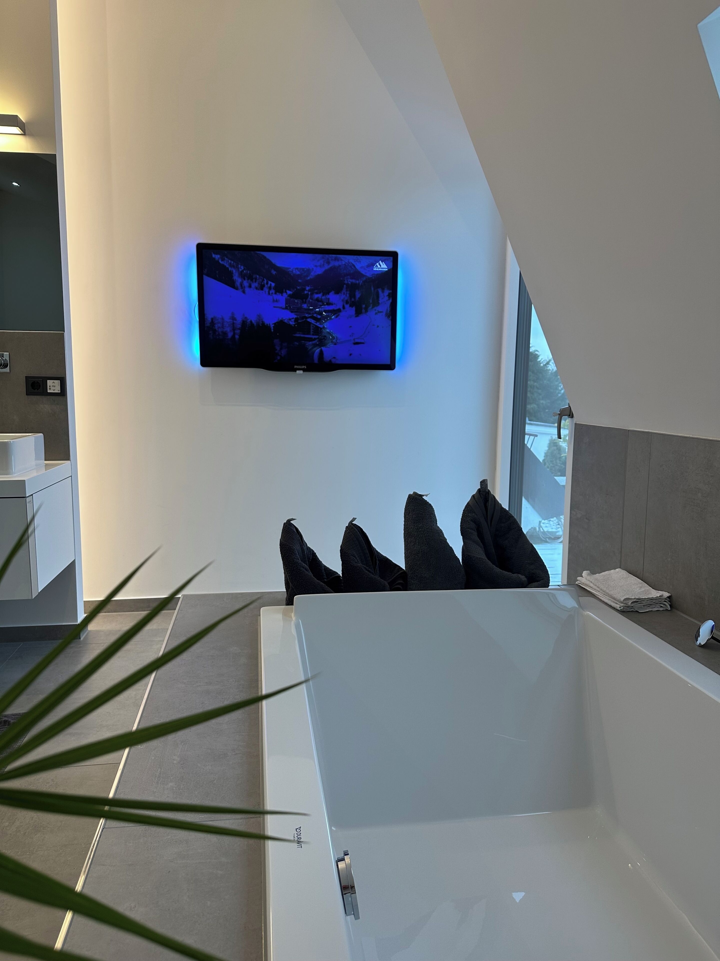 Ein modernes Badezimmer mit einer großzügigen Badewanne, einem angebrachten Fernseher, der eine verschneite Landschaft zeigt, und einem sichtbaren Fenster.