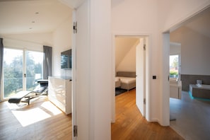 Ein moderner, gut beleuchteter Raum mit Holzboden, einem Liegestuhl, einem weißen Schrank und einem großen Fenster mit Blick auf Bäume.