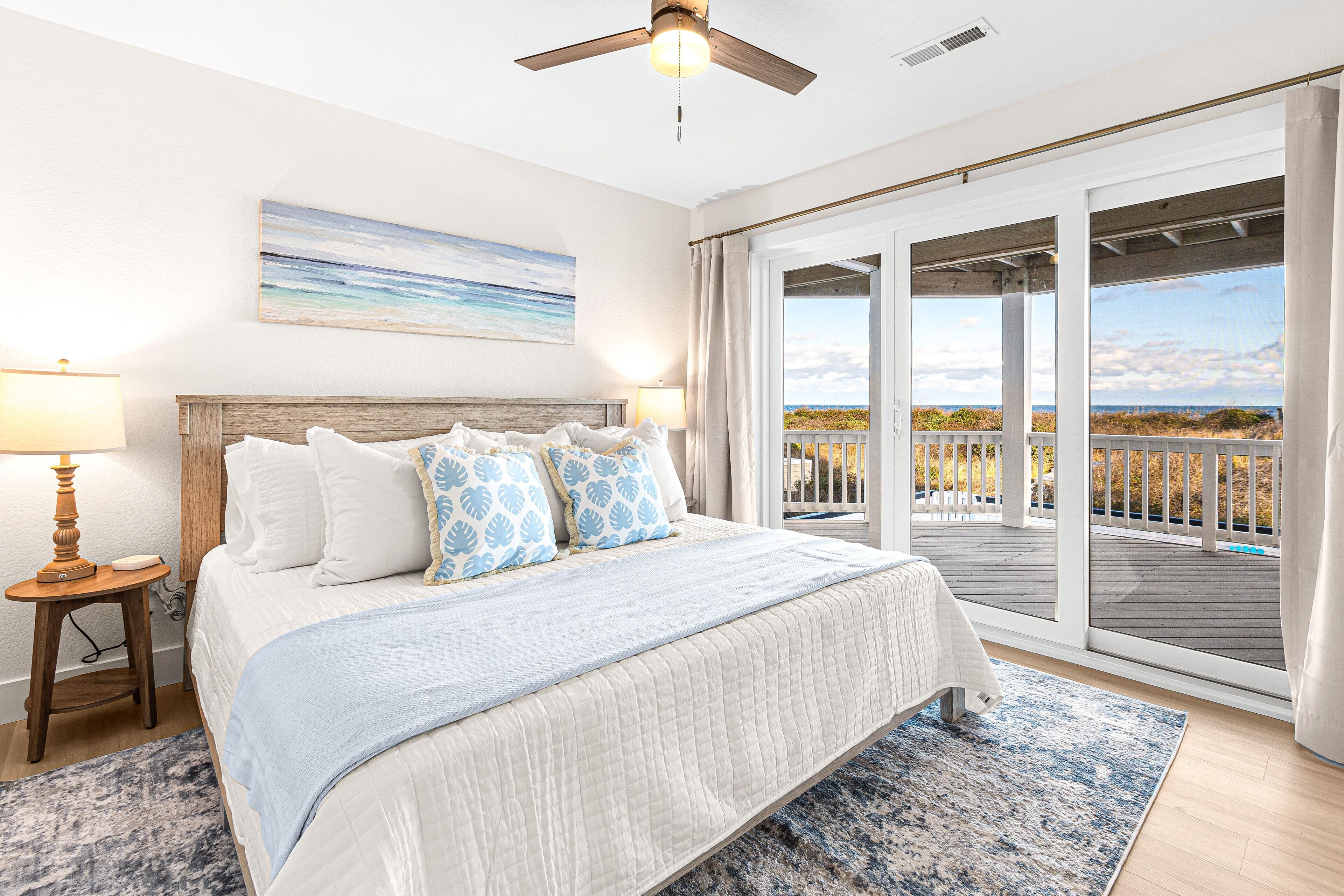 A bedroom with a large bed, a ceiling fan, a nightstand with a lamp, and a view of the beach through the sliding glass doors.