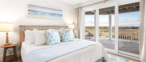 A bedroom with a large bed, a ceiling fan, a nightstand with a lamp, and a view of the beach through the sliding glass doors.