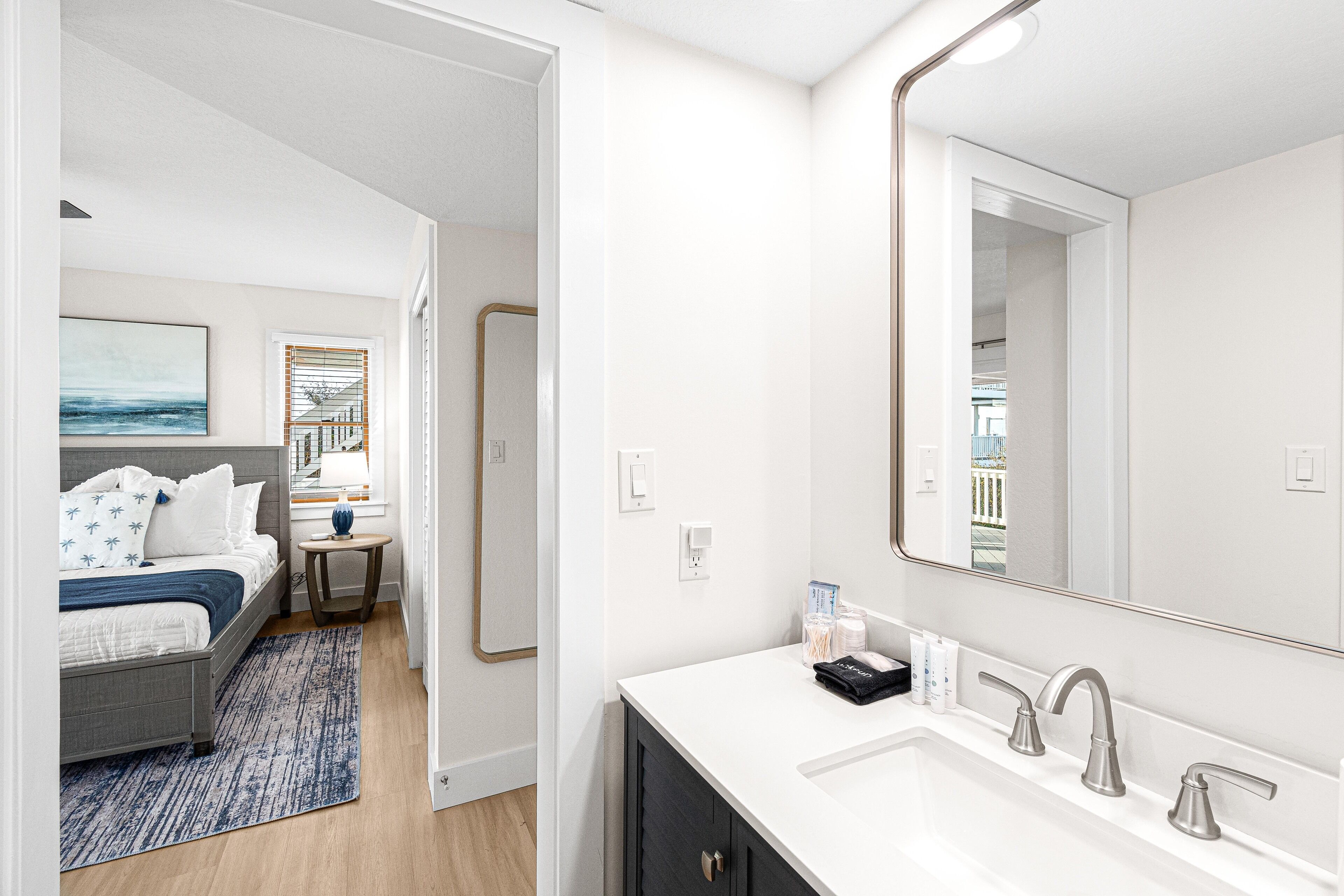 Habitación de hotel moderna con una cama grande, un baño con tocador blanco y un espejo amplio, y vistas de la ciudad a través de la ventana.