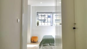 Una habitación con cama, ventana y una pequeña silla naranja.