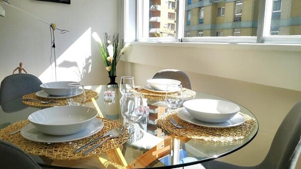 Una mesa de comedor dispuesta con platos, tazones y cubiertos blancos, con vista a un edificio a través de la ventana.