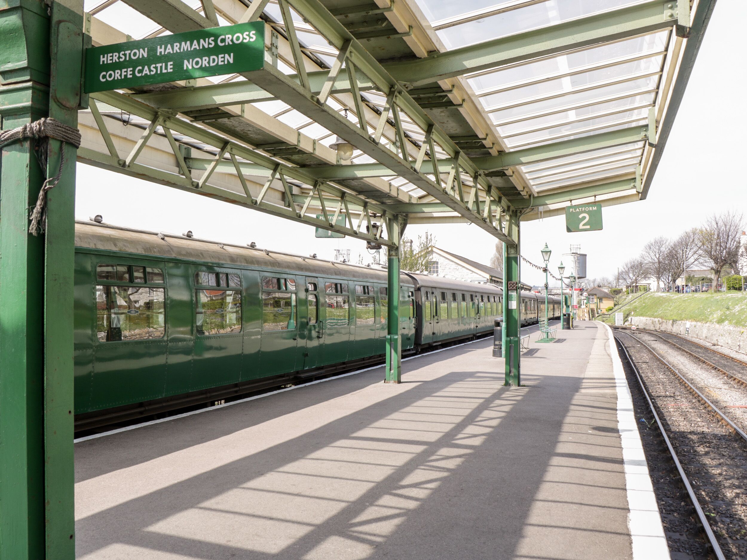 Andén de estación de tren con un tren verde, un refugio y un cartel que indica 'Herston Harmans Cross Corfe Castle Norden'.