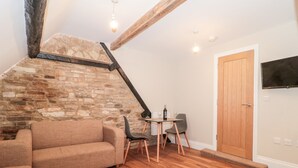 Un salón acogedor con pared de piedra, vigas de madera, un sofá, una mesa con sillas y un televisor montado en la pared.