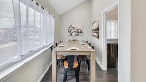 A dining area with a long table set for six, large windows with curtains, and a painting on the wall.