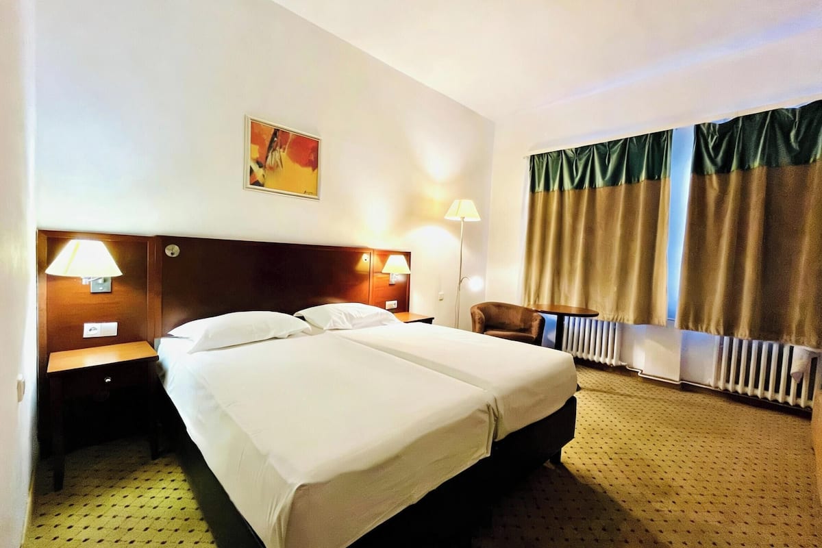 A hotel room with a double bed, two bedside lamps, a headboard, a chair, a radiator, and a window with curtains.