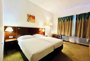 A hotel room with a double bed, two bedside lamps, a headboard, a chair, a radiator, and a window with curtains. - Hotel Braník (Prague)