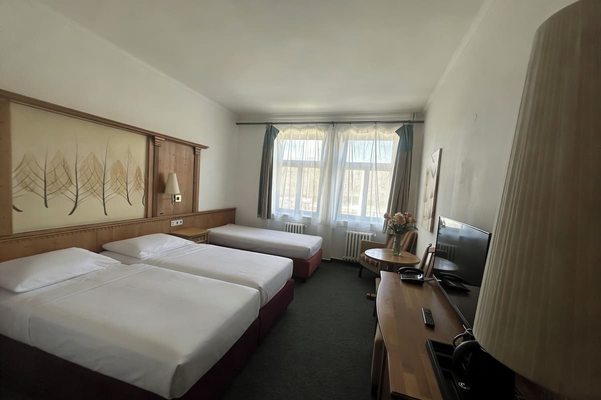 A hotel room with two beds, a desk with a computer, a chair, a radiator, and a window with curtains.