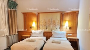 Camera d'albergo con due letti singoli, una testiera in legno decorata con un affresco a motivi floreali, lampade sui comodini e un termosifone.