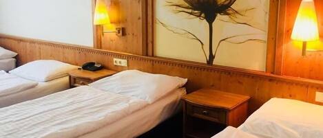 A hotel room with two single beds, wooden headboards, a nightstand with a phone, and a wall-mounted lamp.