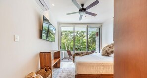 A bedroom with a bed, a ceiling fan, a TV mounted on the wall, and a large window with a view of trees.