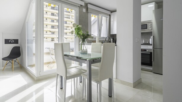 A modern kitchen with stainless steel appliances, a dining area with a glass table and white chairs, and a view of a residential building through large windows. - Atico de dos Dormitorios con Terraza y Litera (Fuengirola)