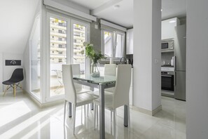 A modern kitchen with stainless steel appliances, a dining area with a glass table and white chairs, and a view of a residential building through large windows.