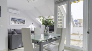 A modern living room with a glass dining table, white chairs, a grey sofa, a framed picture on the wall, and a balcony with a view of buildings. - Atico de dos Dormitorios con Terraza y Litera (Fuengirola)