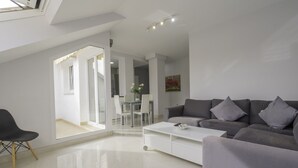 A modern living room with a grey sofa, a white coffee table, and a black chair. There is a glass door leading to a dining area with a white table and chairs. - Atico de dos Dormitorios con Terraza y Litera (Fuengirola)