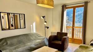 Una sala de estar con sofá, sillón y mesa de centro de madera. Hay una lámpara de pie y un ventanal con vistas a montañas.