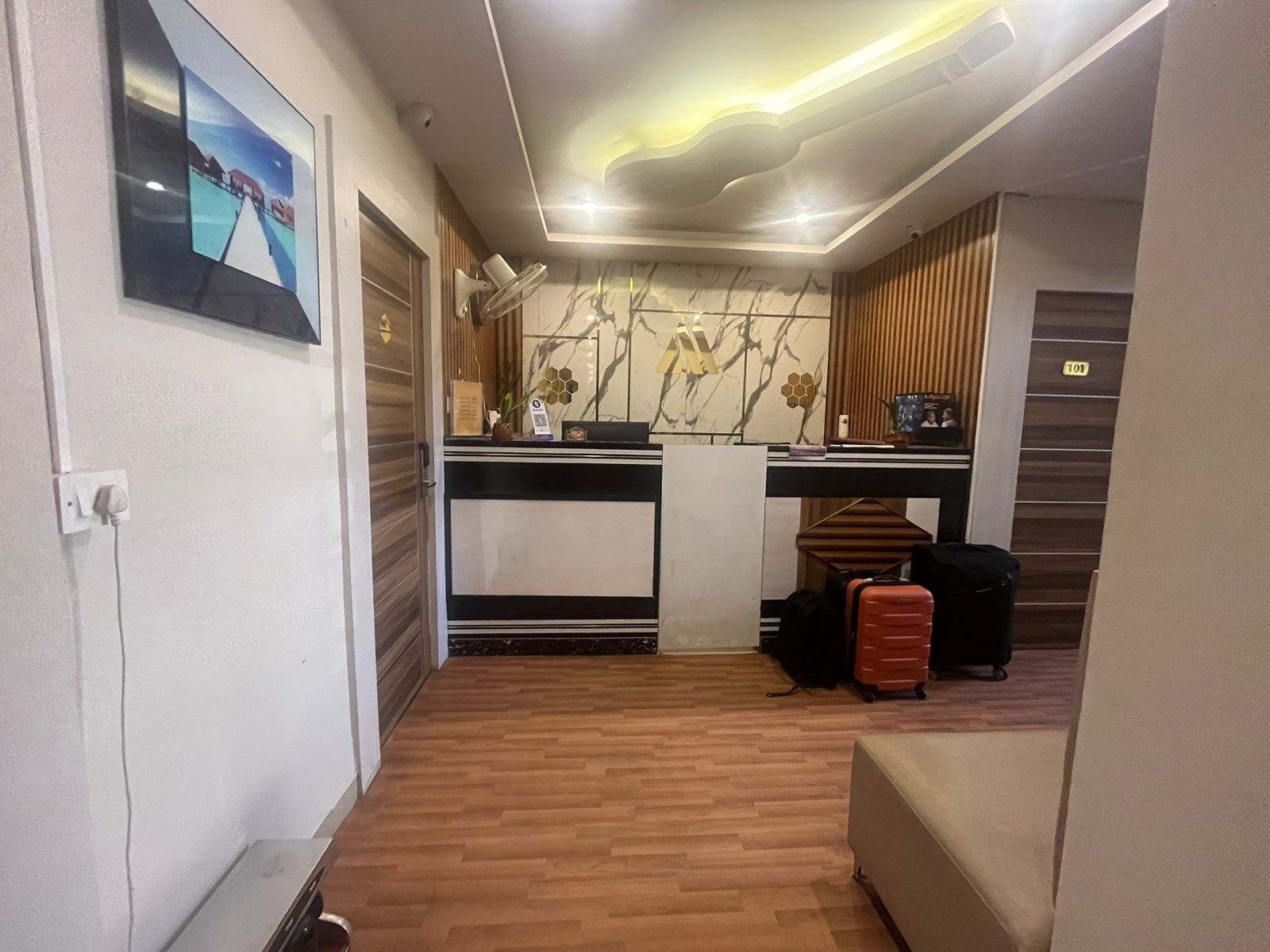 A compact hotel room with a wooden floor, a bed, a desk with a chair, a small table, and a wall-mounted TV.