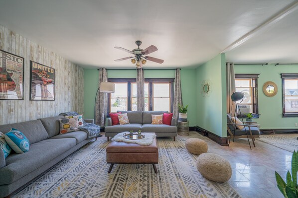 A living room with a grey sofa, a brown ottoman, a patterned rug, a ceiling fan, and a window with curtains.