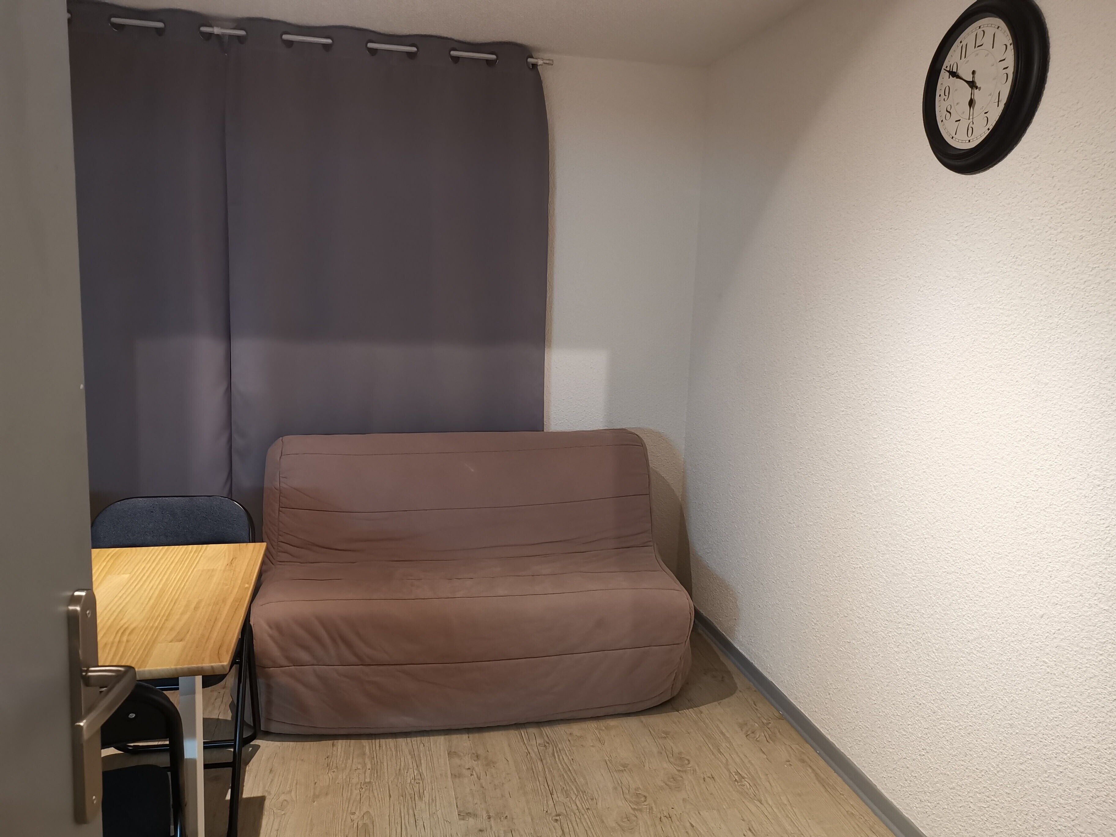 A small room with a brown sofa, a wooden table, a chair, and a wall clock.