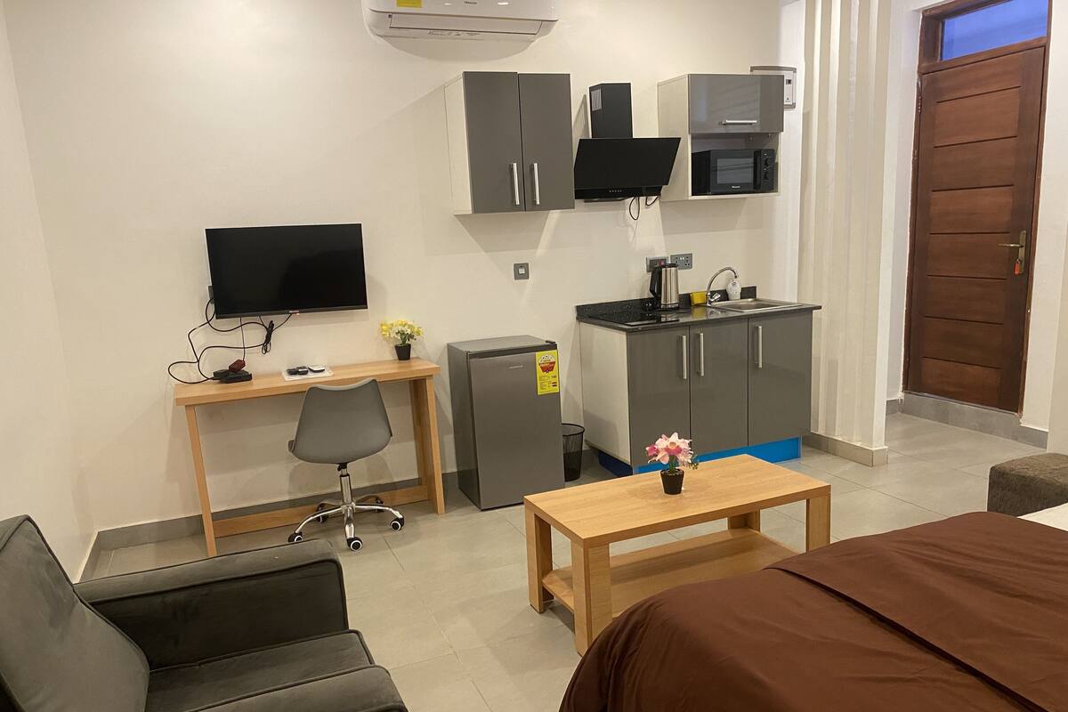 Une chambre d’hôtel de taille réduite, comprenant un lit, un bureau avec une chaise, une kitchenette, un téléviseur et un petit réfrigérateur.