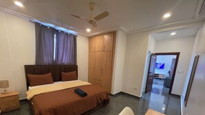 A bedroom with a bed, a wooden wardrobe, a ceiling fan, and a dining area with a table and chairs. - Mayama Residences  (Accra)