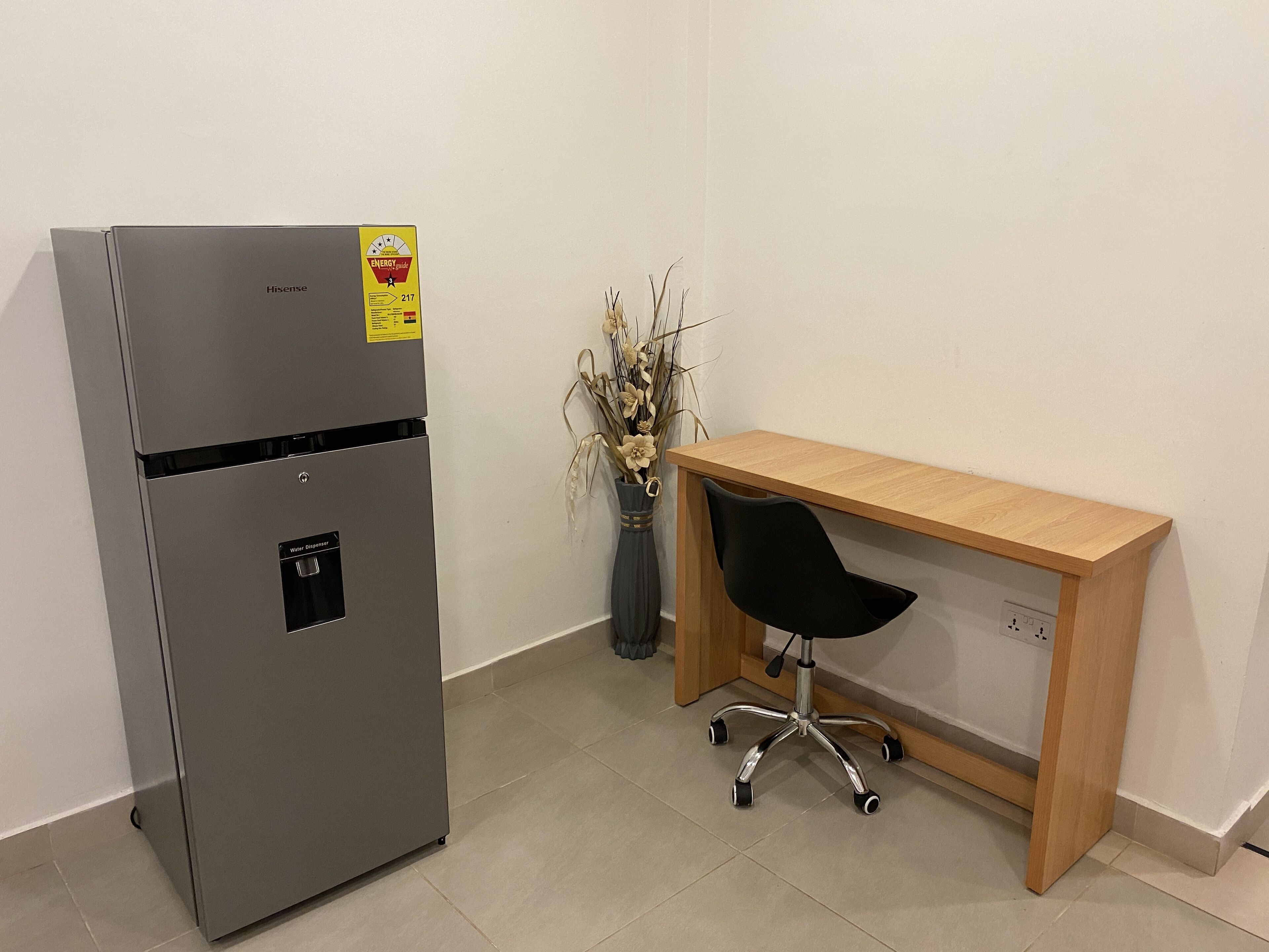 A compact kitchen with a stainless steel refrigerator, a wooden desk with a black office chair, and a vase with dried plants.