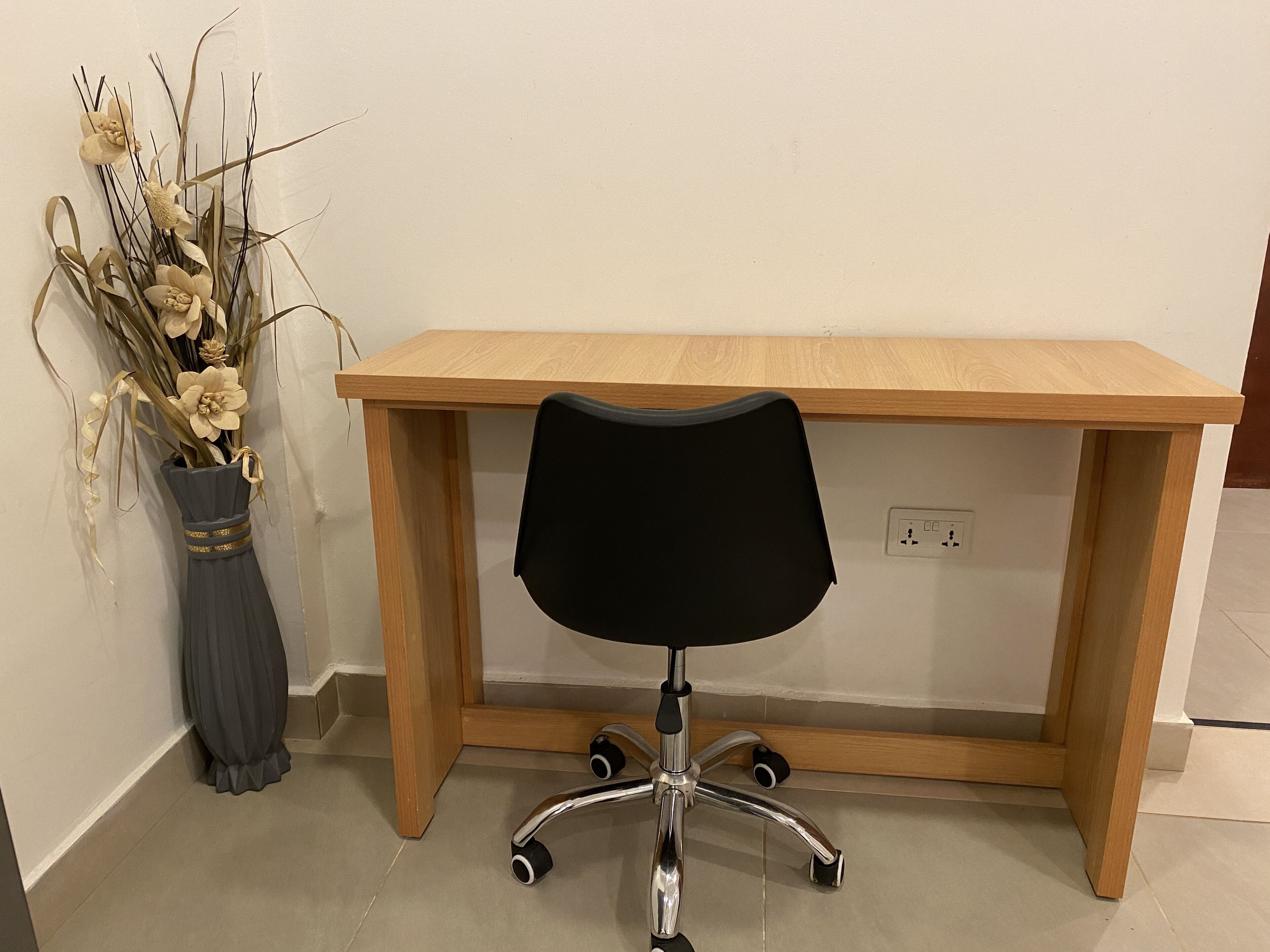 Una scrivania in legno con una sedia da ufficio nera, un vaso con fiori secchi e una presa elettrica sulla parete.