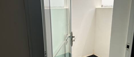 Una esquina de una habitación con una puerta de cristal y una ventana, suelo de baldosas y pared blanca.