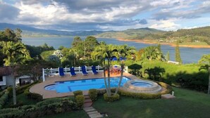 A swimming pool with lounge chairs, surrounded by well-maintained lawns and palm trees, with a view of a lake and distant hills. - Casa Maria, Lagoon Calima (Club Las Anonimos)