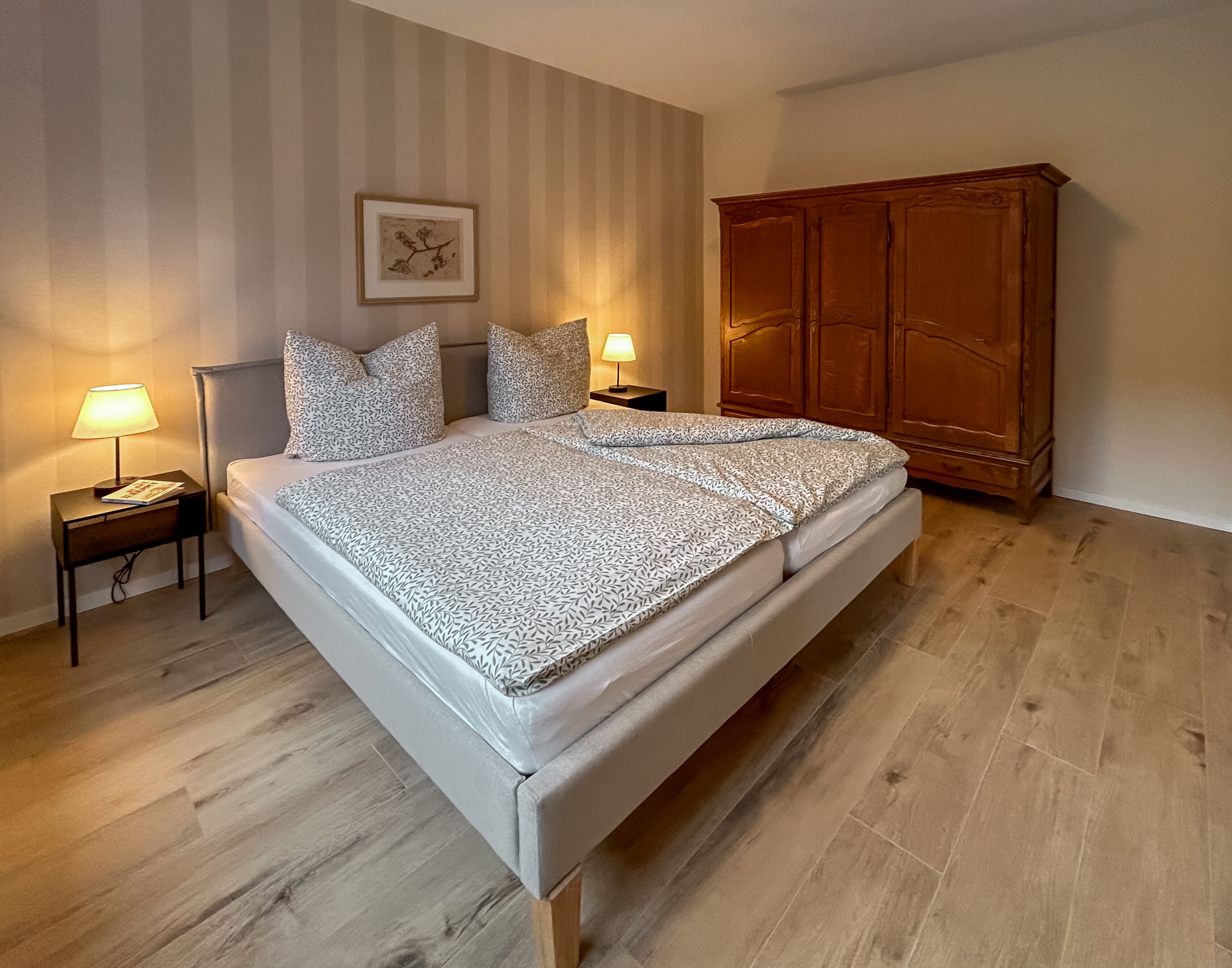 Une chambre bien rangée, avec un lit, deux tables de chevet dotées de lampes, une armoire en bois et un tableau encadré accroché au mur.