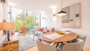 Ein modernes Wohnzimmer mit einem runden Holztisch, grauen Polsterstühlen, einer Couch und einem großen Fenster mit Blick auf Grünflächen.
