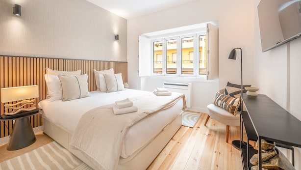 Ein modernes Schlafzimmer mit einem großen Bett, Nachttischen, einem Schreibtisch und einem Fenster mit Blick auf Gebäude.