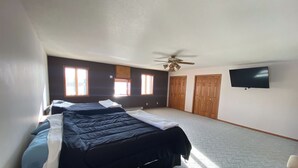 Un dormitorio con una cama grande, ventilador de techo, televisor y dos ventanas.
