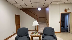 Una habitación con piso alfombrado, dos sillones, una mesita con una lámpara, una escalera y una puerta que da a otra habitación.