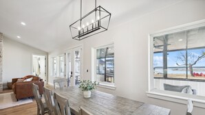 Un comedor amplio con una mesa y sillas de madera, una lámpara colgante y ventanas grandes con vista a la playa.